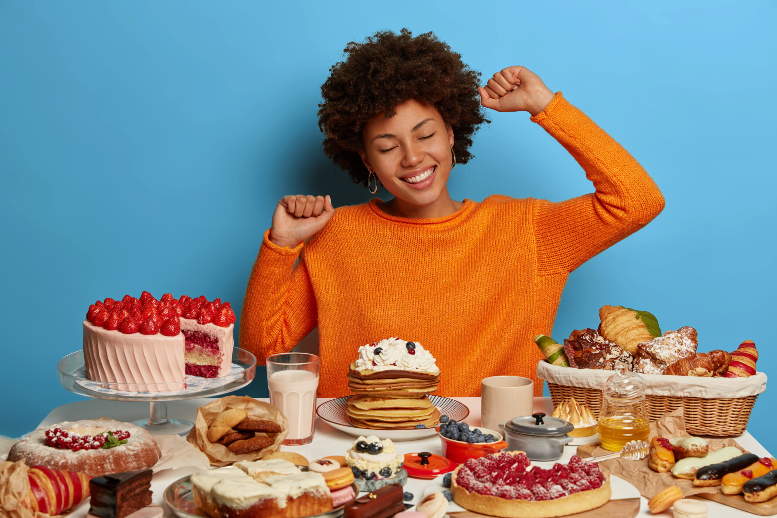 Sweet tooth, temptation by delicious food. Pretty woman with Afro hairstyle has good mood after eating tasty delicacy, stretches at table with confectionery. Laziness, overeating and gluttony