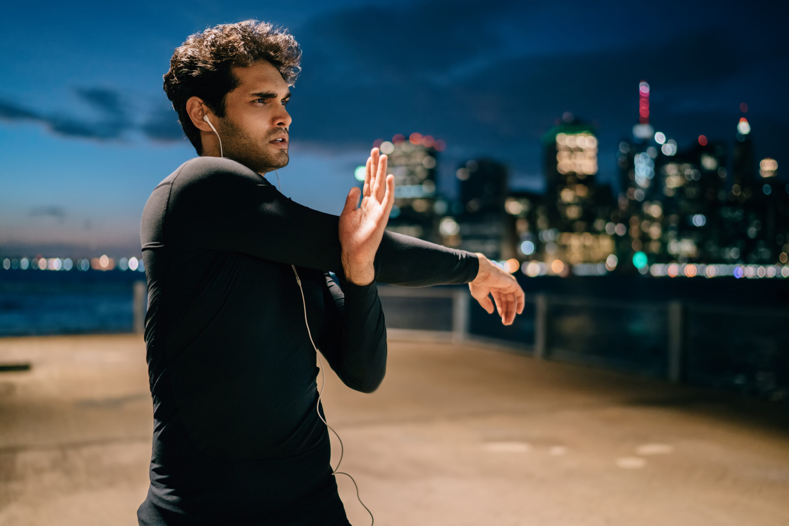 Sport motivated man listening music from playlist in earphones and doing stretching hands during evening workout in urban setting.Sportsman in active wear warming up before training in night city