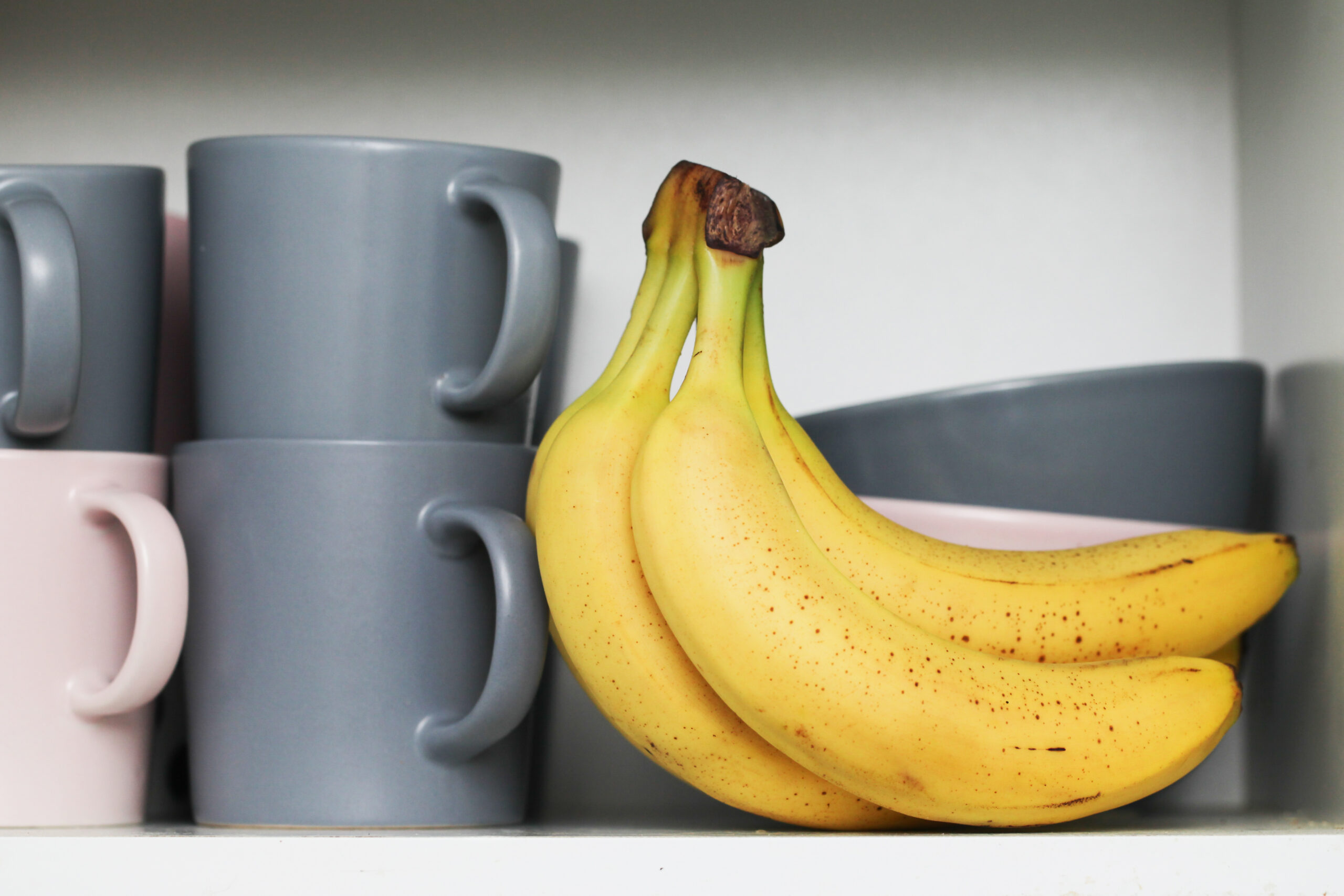 Bananas in kitchen cabinet. Coffee mugs. Tea cups. Milk soup bowls. Yellow fruits in kitchen. Open door kitchen cabinet with hidden bananas inside. Ripe bananas with small brown stains.